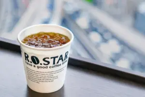 5943a34d122ee24017ef75436a27f9a5 | コーヒーステーション Iced coffee in a white takeout cup with bold black ROSTAR lettering on a wooden counter with a blurred city background