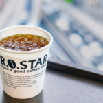 Paper cup of iced coffee labeled ROSTAR on a wooden ledge with a blurred cityscape in the background