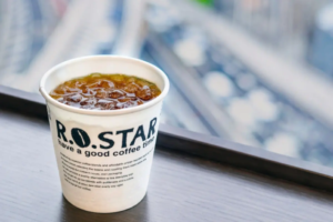 Paper cup of iced coffee labeled ROSTAR on a wooden ledge with a blurred cityscape in the background