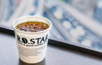 Paper cup of iced coffee labeled ROSTAR on a wooden ledge with a blurred cityscape in the background