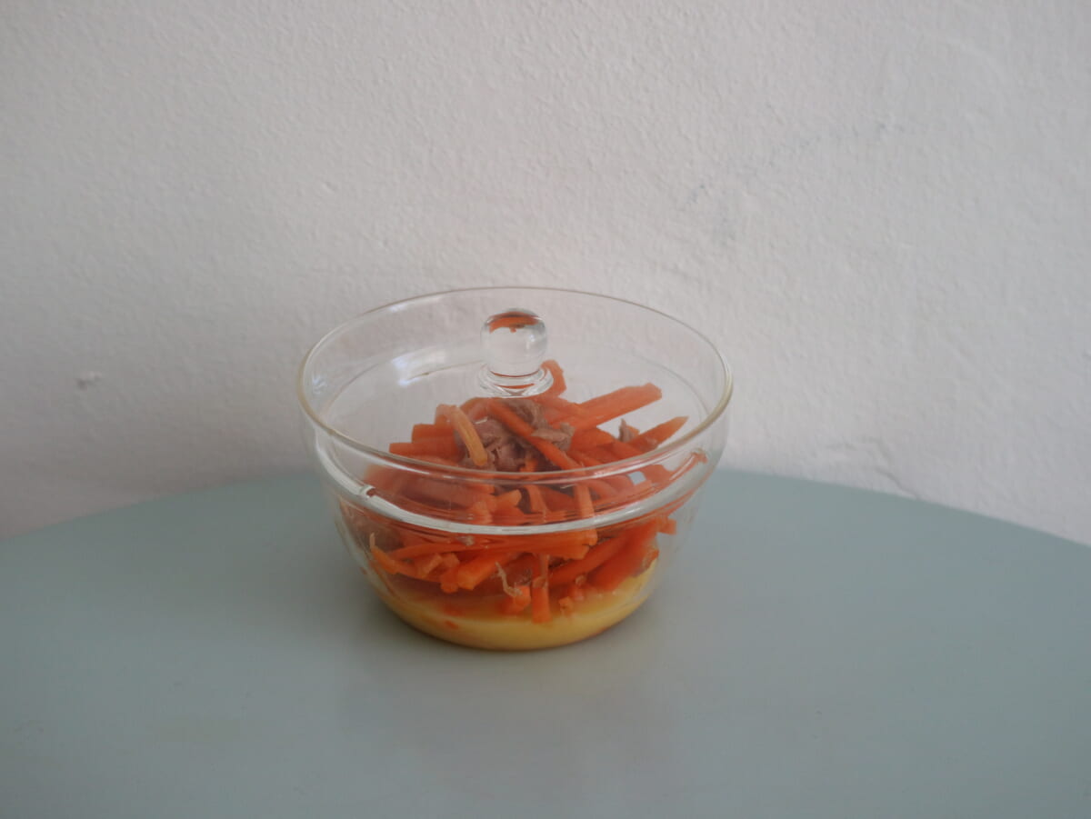 Glass bowl with shredded orange carrots resting on a yellow base placed on a pale blue table against a white wall