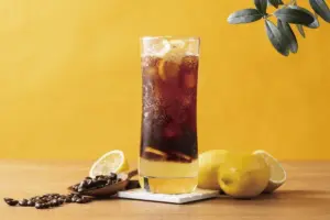 6f39afe8557a6ddaee89a08a687a2802 | コーヒーステーション Tall glass of iced coffee with lemon slices and ice on a wooden table with coffee beans and lemons nearby