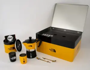 The North Face camping coffee kit with a moka pot cups and spoons laid out beside a yellow storage box with its lid open