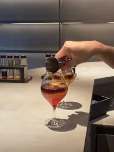 Hand pouring an amber cocktail from a jigger into a stemmed glass at a bar counter