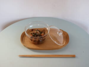 Glass bowl with cooked mushrooms on a wooden tray with lid beside chopsticks on a pale blue table