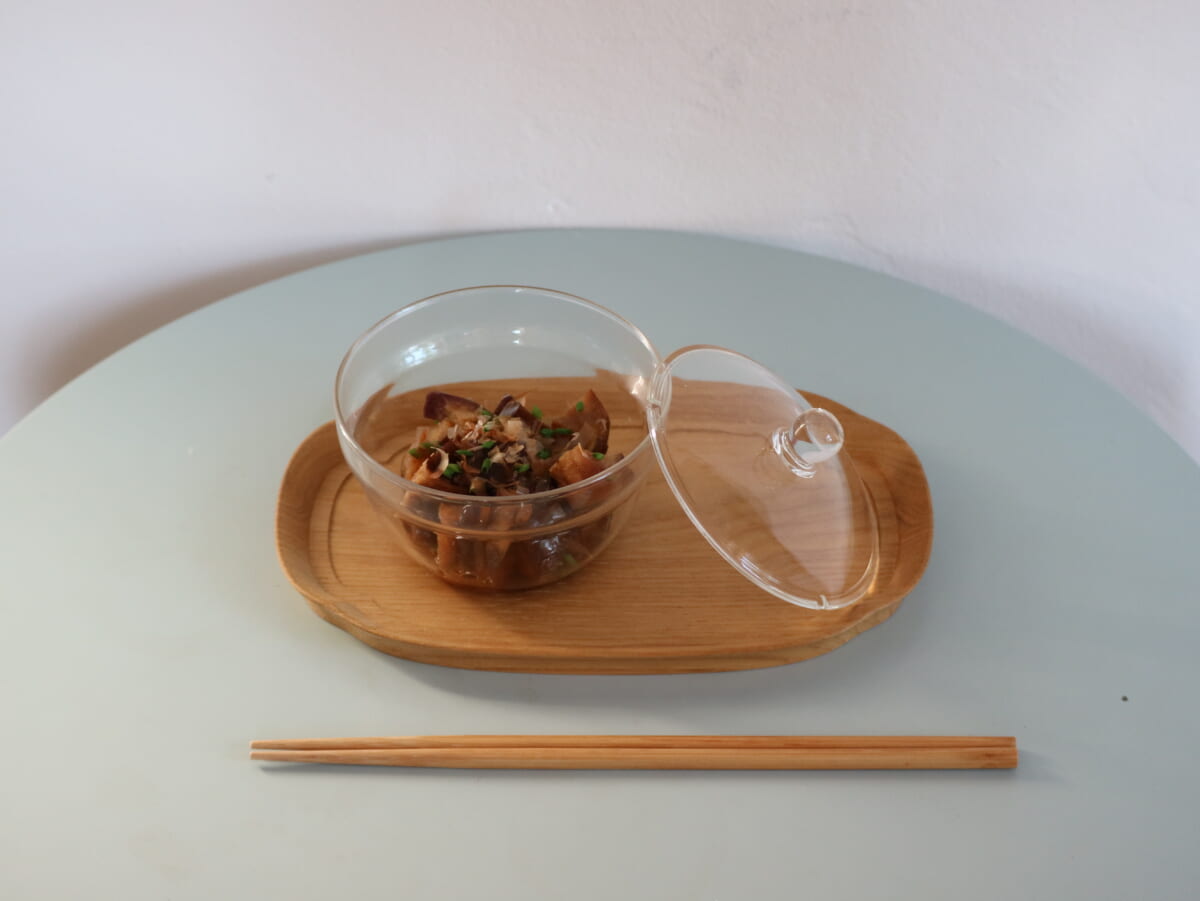 Glass bowl with cooked mushrooms on a wooden tray with lid beside chopsticks on a pale blue table