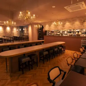 Restaurant interior with a long wooden bar high stools and chandeliers open kitchen in the background and brick walls