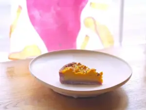 7e760a7a6608702fe5d8dd6e57ef53f5 | コーヒーステーション A slice of two tone tart on a white plate with a purple crust and yellow creamy filling on a wooden table by a bright blurred background