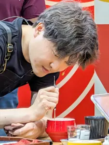 Craftsman leaning over a red lacquer bowl using a thin tool to carve or decorate its interior