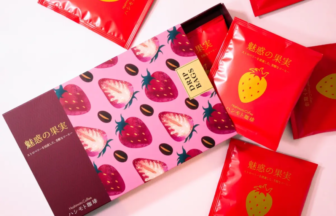 Flat lay of a pink box with strawberry pattern, surrounded by red packets with yellow strawberry illustrations, suggesting strawberry-flavored coffee gifts.
