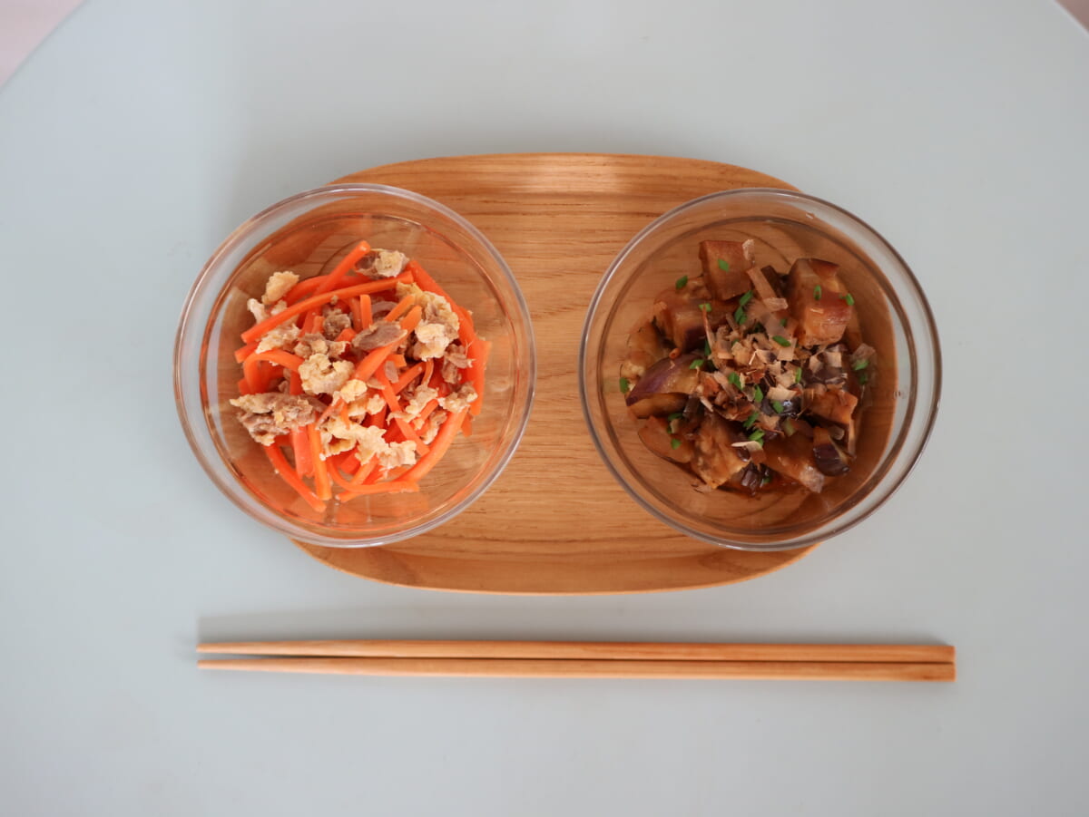 Two glass bowls on a wooden tray with chopsticks beneath left bowl has carrot salad with crumbled topping right bowl contains a braised eggplant dish with herbs and toppings