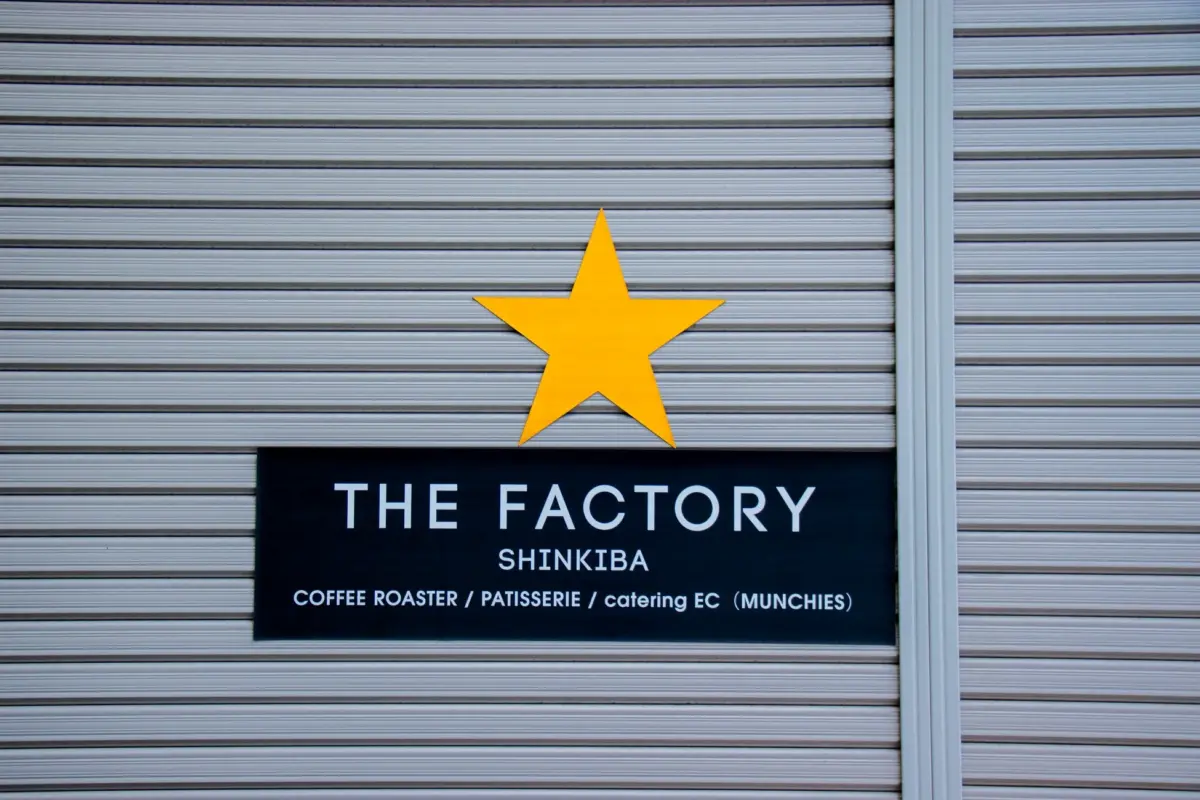 シャッターに設置された「THE FACTORY SHINKIBA」の看板と、その上にある大きな黄色の星の画像です。コーヒーロースター、パティスリー、ケータリングECを扱う店舗であることが示されています。