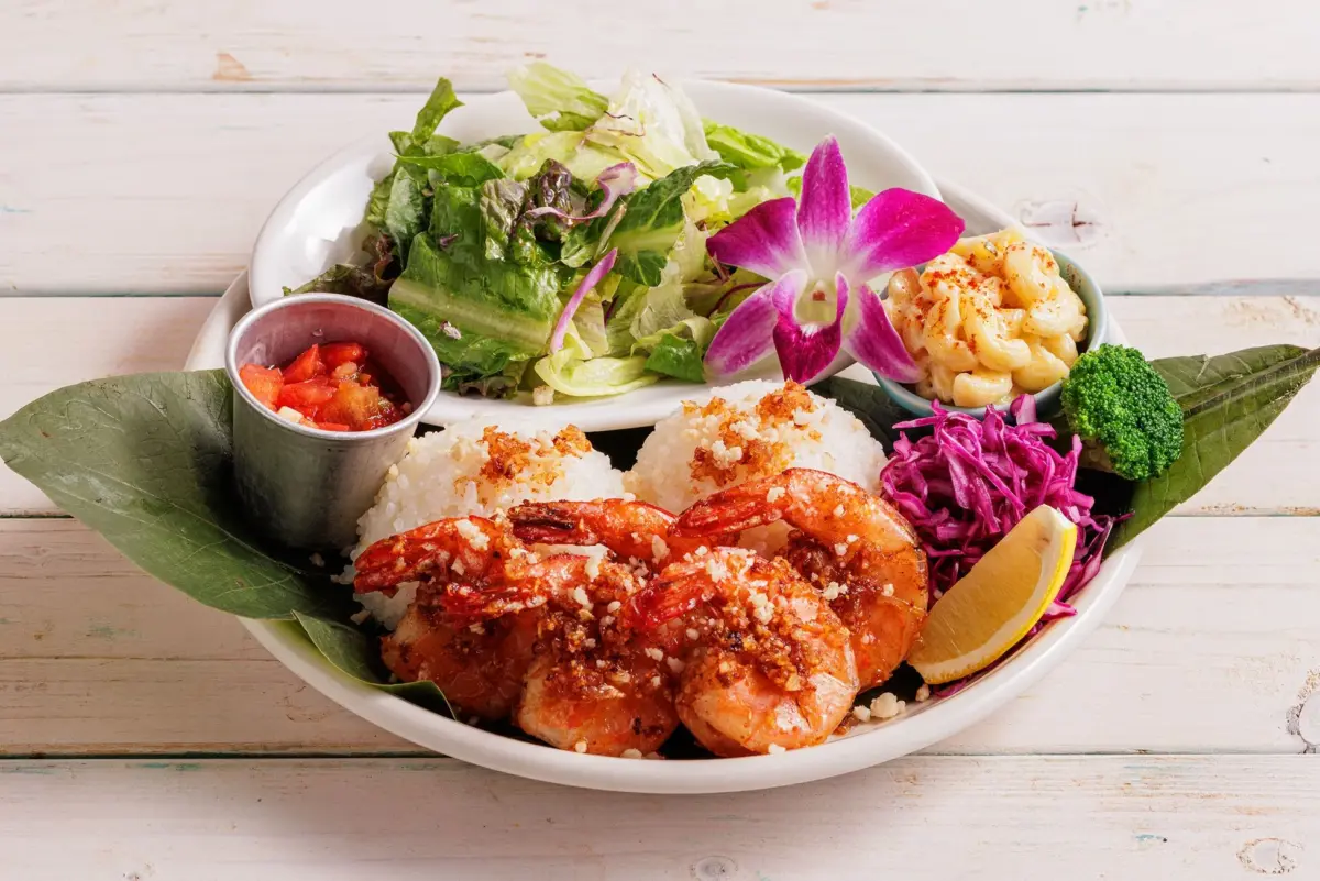 白い皿に盛り付けられたハワイアンガーリックシュリンププレートランチ。香ばしいガーリックシュリンプをメインに、白いご飯、フレッシュなサラダ、マカロニサラダ、ブロッコリーが添えられ、鮮やかな紫色の花とレモンが彩りを添えています。