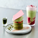 Stack of matcha pastries with a pink chocolate shard cream and strawberry slices on a plate a small green sauce shot beside it and a layered matcha pink drink in the background