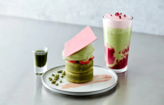 Stack of matcha pastries with a pink chocolate shard, cream, and strawberry slices on a plate; a small green sauce shot beside it and a layered matcha-pink drink in the background.