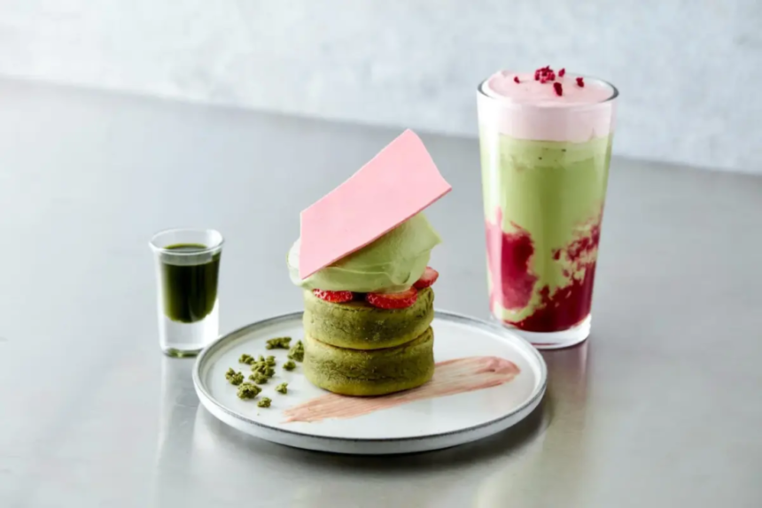 Stack of matcha pastries with a pink chocolate shard cream and strawberry slices on a plate a small green sauce shot beside it and a layered matcha pink drink in the background