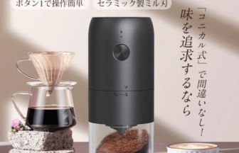 Black electric coffee grinder with glass chamber, surrounded by a pour-over setup and a white cup of coffee on a light background
