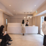 Minimalist cafe reception with a white counter staff talking to a customer and a seated person on the left sipping from a cup