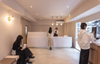 Minimalist cafe reception with a white counter, staff talking to a customer, and a seated person on the left sipping from a cup.