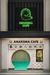 Front of ANAKUMA CAFE storefront featuring a circular arched doorway with teal interior floor visible and a neon green cat logo above with the cafe name on a signboard
