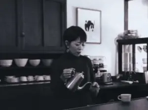 982567e53101d3eb9c0a569069e4e058 | コーヒーステーション Woman at a cafe counter pours hot water from a kettle into a cup with shelves of dishes behind her