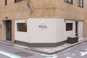 99045c29faf5eda4bda633ee8629234d | コーヒーステーション Corner cafe storefront with a white facade on a brick building sign reads Daily Tea Coffee beside two small windows and a bench on the sidewalk