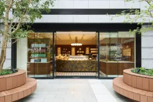 Modern storefront with glass doors opening to a bakery counter inside surrounded by planters and trees outside