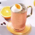Copper mug of iced drink topped with whipped cream and orange jam on a yellow coaster orange half and whole orange in the background with a café logo in the top left corner