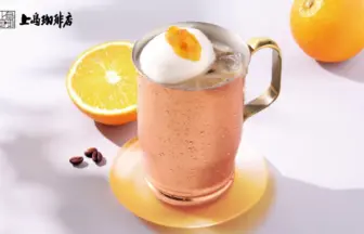 Copper mug of iced drink topped with whipped cream and orange jam on a yellow coaster orange half and whole orange in the background with a café logo in the top left corner