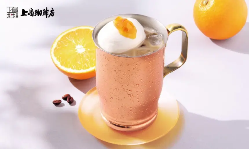 Copper mug of iced drink topped with whipped cream and orange jam on a yellow coaster orange half and whole orange in the background with a café logo in the top left corner
