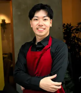 Smiling staff member in a red apron with arms crossed indoors facing the camera