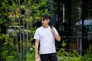 Man in a white t shirt and sunglasses stands among green shrubs outside a glass front building with a jacket over his shoulder