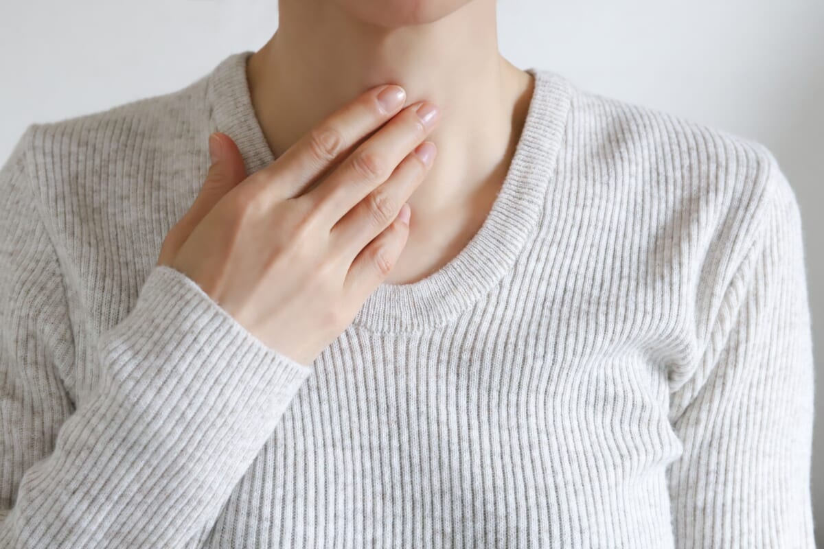 Person in a light gray ribbed sweater touching their throat with one hand suggesting a sore throat or throat discomfort