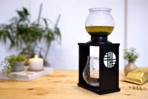 Siphon coffee maker on a black stand with a glass globe containing yellow liquid on a wooden table with a candle and plants blurred in the background