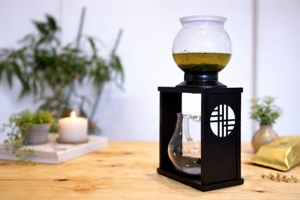 Siphon coffee maker on a black stand with a glass globe containing yellow liquid on a wooden table with a candle and plants blurred in the background