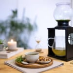 Cozy tea setup on a wooden table glass kettle inside a black lantern a mug of tea bowl of nuts candle hourglass and plants