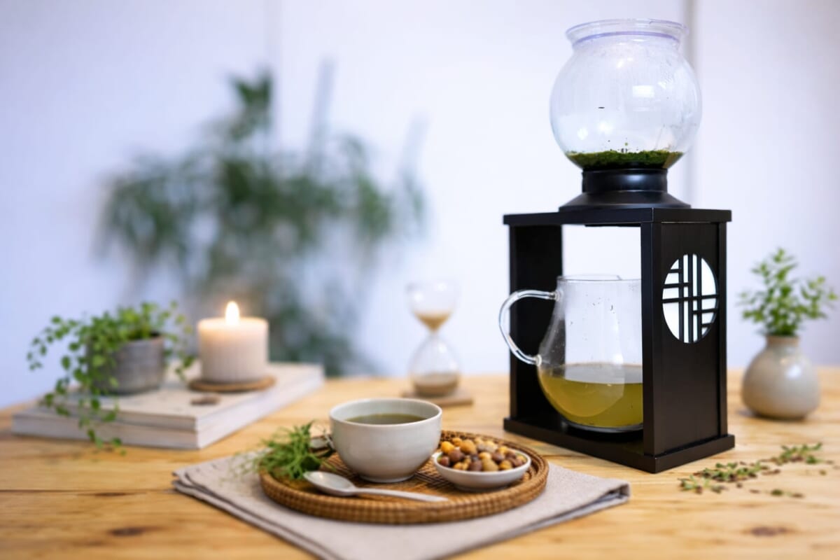 Cozy tea setup on a wooden table glass kettle inside a black lantern a mug of tea bowl of nuts candle hourglass and plants
