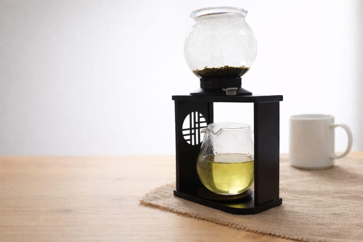Glass siphon coffee maker on a black stand with a yellow green liquid in the lower bulb mug in the background on a wooden table