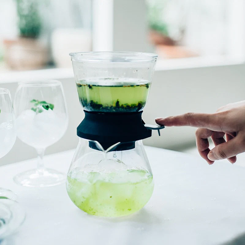 Person pressing a lever on a glass tea siphon brewing bright green tea with two empty wine glasses beside it