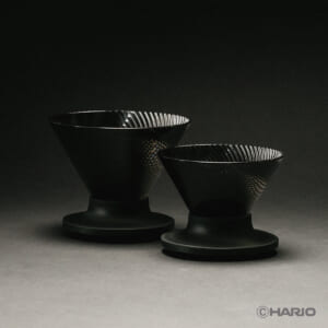 VDN57146a4e8ecc41dc9ca3b7c4e7b0aa14 | コーヒーステーション Two black Hario V60 coffee drippers with ribbed interiors resting on circular bases in a dark studio setting