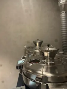 a22386f7139dc230cec8e3c87ca4b461 | コーヒーステーション Industrial coffee roasting machine with stainless steel drum and funnel chutes vent hose visible on the right side setup in a lab like room