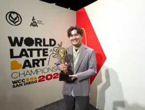 Smiling man in a striped blazer holding a latte art championship trophy in front of a World Latte Art Championship banner
