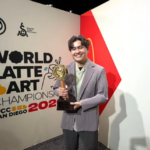 Person smiling and holding a trophy at the World Latte Art Championship 2022 in San Diego standing in front of a wall with the event sign
