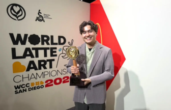 Person smiling and holding a trophy at the World Latte Art Championship 2022 in San Diego standing in front of a wall with the event sign