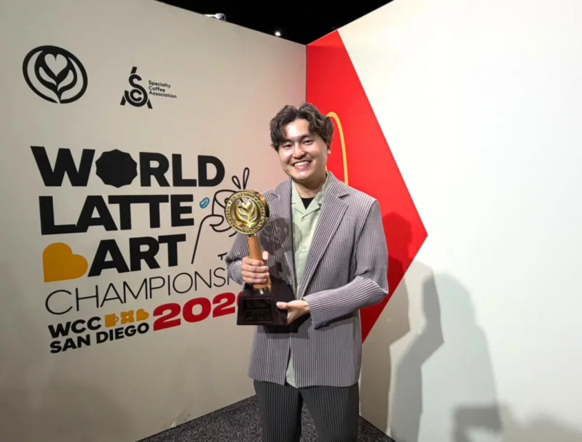 Person smiling and holding a trophy at the World Latte Art Championship 2022 in San Diego standing in front of a wall with the event sign