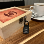 Wooden puzzle box secured with a four digit combo padlock on a wooden table beside a white coffee cup and saucer with a spoon