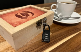 Wooden puzzle box secured with a four digit combo padlock on a wooden table beside a white coffee cup and saucer with a spoon