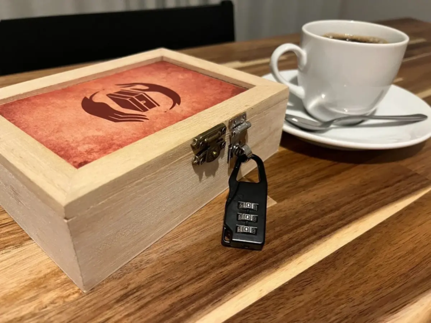 Wooden puzzle box secured with a four digit combo padlock on a wooden table beside a white coffee cup and saucer with a spoon