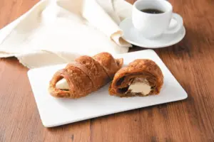Two cream filled croissants sliced open on a white plate with a cup of coffee and a napkin on a wooden table