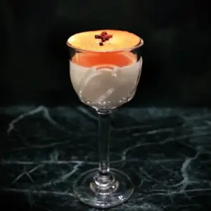 Cocktail in a stemmed glass with a foamy orange top and a small garnish served on a dark marble surface